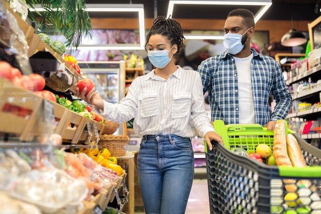 Familienlebensmitteleinkaufen. Schwarzes Paar, das Gemüse und Obst zusammen mit Shop-Trolley-Wagen im Supermarkt-Lebensmittelgeschäft kauft und eine chirurgische Maske trägt. Kunden, die sich für gesundes Essen entscheiden.