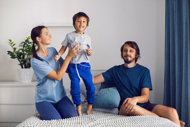 Familienmutter, Vater sitzen und Sohn springen auf weißes Bett im Schlafzimmer am Fenster