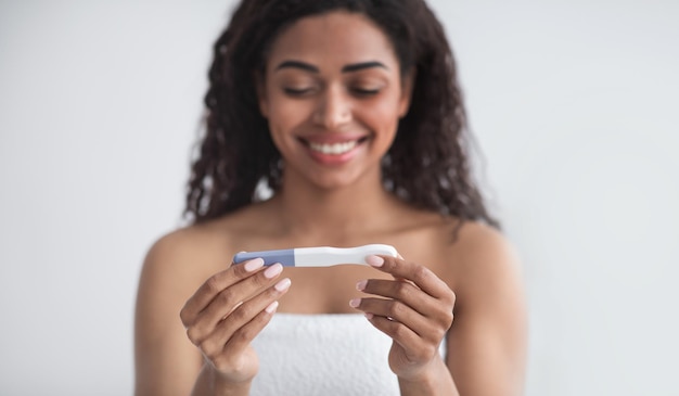 Familienplanung, Eisprung, tolle Neuigkeiten, Baby erwartet. Lächelnde junge Afroamerikanerin betrachtet positiven Schwangerschaftstest, auf grauem Wandhintergrund, selektiver Fokus, Panorama, leerer Raum