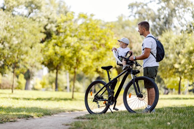 Familienradfahren. Ein Vater und ein Sohn machen an einem sonnigen Sommertag eine Pause vom Radfahren in einem grünen Park. Ein Kleinkind mit Mütze sitzt in einem Fahrradkorb, während Papa neben ihm steht. Seitenansicht