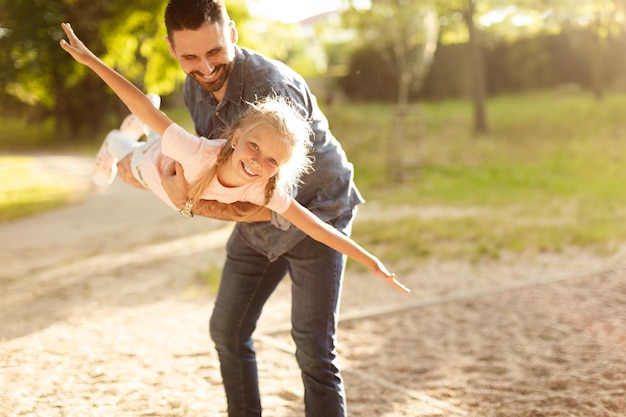 Familientag im Freien, glücklicher Vater mittleren Alters, der die furchtlose Tochter in den Händen hält, Mädchen, das vorgibt zu fliegen