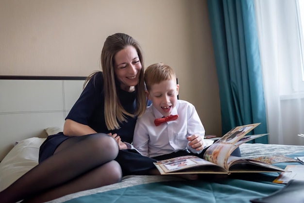 Familienurlaub Fröhliche junge Mutter mit einem rothaarigen Jungensohn, der zu Hause ein gefaltetes Album aus dem Kindergarten auf dem Bett anschaut