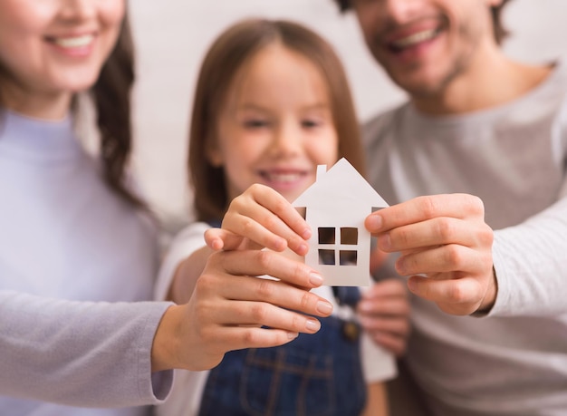 Familienwohnungs- und Annahmekonzept. Ausgeschnittenes Papierhaus in den Händen einer glücklichen dreiköpfigen Familie mit kleiner Tochter, selektiver Fokus