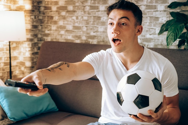 Fan boy viendo fútbol en la televisión sosteniendo un balón de fútbol decepcionado por el gol perdido de su equipo