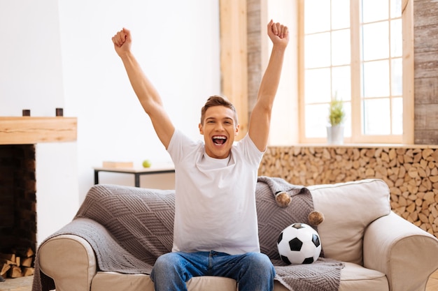 Foto fanático del fútbol. adolescente bien parecido, encantado, bien formado, sonriendo y levantando los brazos mientras está sentado en el sofá y con un balón de fútbol cerca de él.