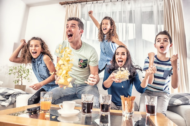 Fanáticos delirantes animando deportes, mientras una familia de cinco miembros ven fútbol o un partido de hockey sobre hielo en la televisión de su casa, gritando de alegría.