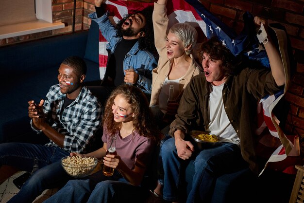 Los fanáticos del fútbol de emocionados amigos americanos multiculturales reunidos en casa en una habitación oscura viendo en la televisión el juego deportivo se siente eufórico esperando gol, pasatiempo, espectadores de torneos, concepto de apuestas deportivas