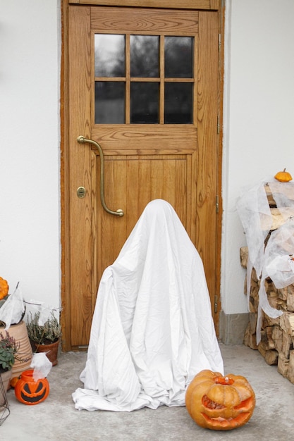 Fantasma espeluznante de pie en el frente de la casa con Jack o linterna calabaza telaraña arañas murciélagos y velas Niño disfrazado de fantasma en la decoración frontal de Halloween Feliz Halloween Truco o trato