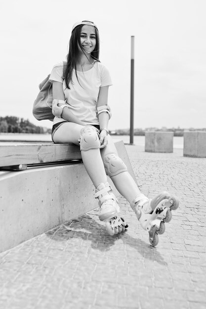 Fantástica mujer joven con ropa informal y gorra sentada en el banco en el parque de patinaje con patines en una foto en blanco y negro