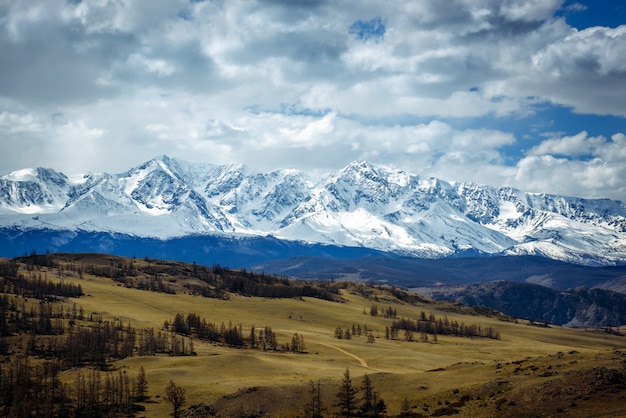 Fantástica paisagem de montanha. Montanhas rochosas com picos nevados, colinas cobertas com grama na cena alpina em um dia brilhante de outono com céu azul e nuvens. Vista das estepes e montanhas cobertas de neve