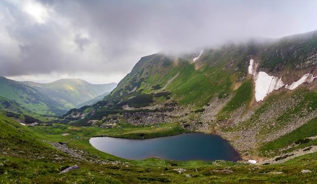 Fantástica vista amplia y pacífica del lago azul en el valle verde y pequeñas tiendas turísticas en la montaña rocosa con parches de nieve cubiertos de nubes bajas. Belleza de la naturaleza, el turismo y el concepto de viaje.