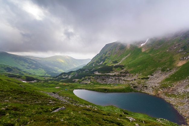 Fantástica vista amplia y pacífica del lago azul en el valle verde y pequeñas tiendas turísticas en la montaña rocosa con parches de nieve cubiertos de nubes bajas. Belleza de la naturaleza, el turismo y el concepto de viaje.