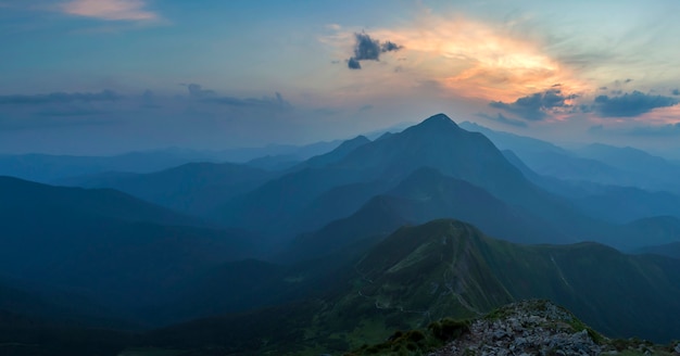 Fantástico amanecer o atardecer sobre la cresta verde de la montaña cubierta de densa niebla azul. Sol anaranjado brillante que levanta en cielo nublado suave sobre horizonte distante. Belleza de la naturaleza, el turismo y el concepto de viaje.