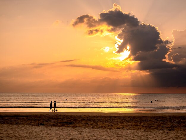 Fantástico céu de pôr-do-sol com belos raios de sol atrás da nuvem sobre o fundo de água do mar com silhueta de pessoas na costa Praia de areia de verão à noite Nascer do sol de manhã cedo ou crepúsculo paisagem marinha