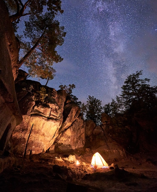 Fantástico paisaje en la noche de verano. brillantemente ardiendo una pequeña fogata en el cañón en medio de una gran formación de rocas empinadas bajo un claro cielo estrellado oscuro. concepto de turismo, seguridad, escalada, senderismo y viajes.