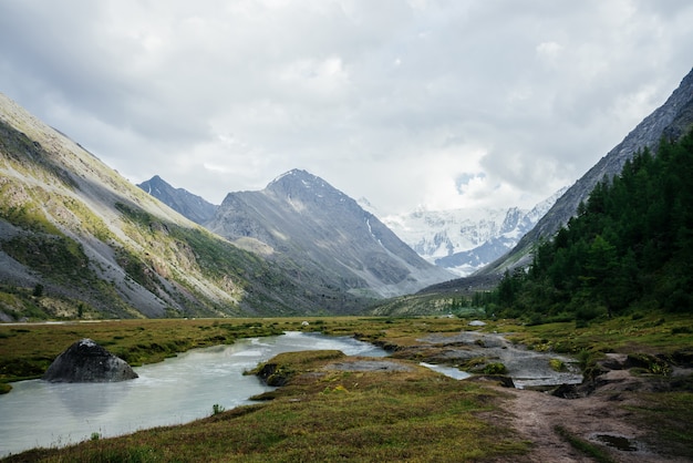 Fantastische alpine Aussicht auf Bergsee und große Gletscher unter düsterem Himmel. Dunkle atmosphärische Hochlandlandschaft mit hohen schneebedeckten Bergen und großen Felsen. Wunderbare Berglandschaft bei bewölktem Wetter.