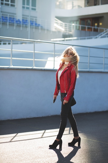 Fantastische blonde Frau in roter Jacke, die mit flatternden Haaren die Straße entlang geht