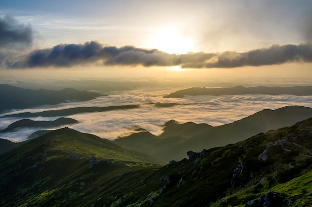 Fantastischer Blick auf das Gebirgstal, bedeckt mit niedrigen weißen geschwollenen Schneewolken, die sich bis zum nebligen Horizont unter hellem Morgenhimmel mit hellem orangefarbenem Schein bei Sonnenaufgang erstrecken. Schönheit der Natur Konzept.