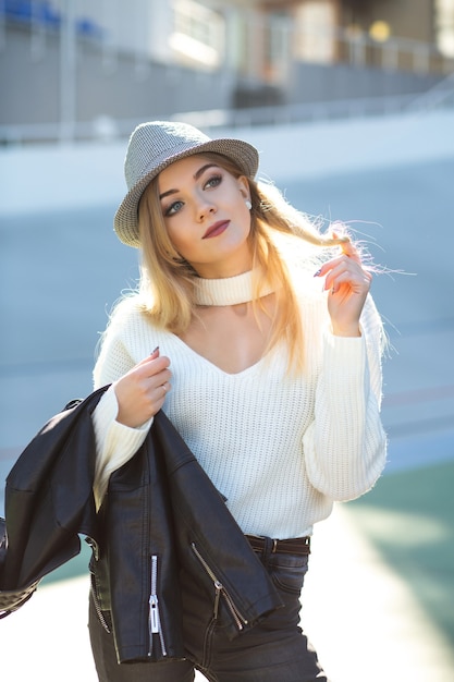 Fantastisches blondes Model mit Hut und Strickpullover, das am sonnigen Tag posiert