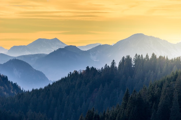 Farbenfroher Sonnenuntergang in den bayerischen Alpen. Blaue Berggipfel, grüner Wald und heller Himmel.