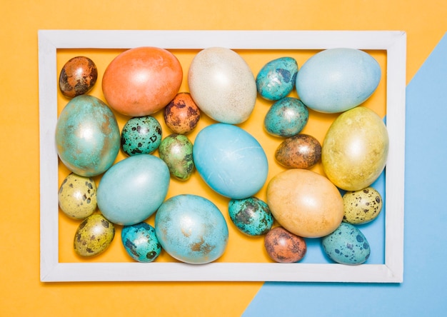 Farbige Ostereier in einem weißen Holzrahmen auf farbigem Hintergrund. Frohe Ostern!