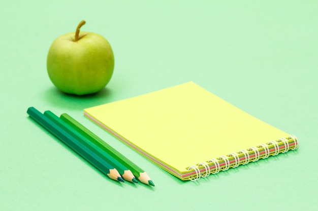 Farbstifte, Notizbuch und Apfel auf grünem Hintergrund.