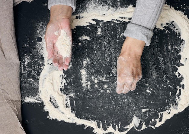 Farinha de trigo branca peneirada em uma mesa preta e duas mãos femininas vista superior cozinhando em casa