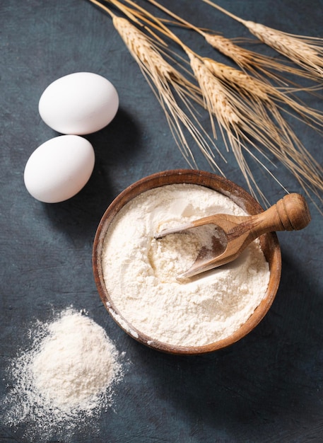 Farinha de trigo em uma tigela de madeira com uma colher em um fundo azul escuro com ovos e galhos secos de trigo Ingredientes orgânicos para assar Conceito de comida saudável caseira