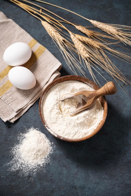 Farinha de trigo numa tigela de madeira com uma colher e ovos sobre um fundo escuro Farinha orgânica natural para assar em casa