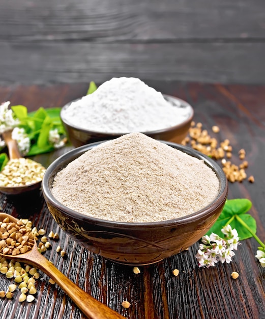 Farinha de trigo sarraceno de cereais verdes e marrons em duas tigelas, colheres com sêmolas, flores e folhas no fundo de uma placa de madeira escura