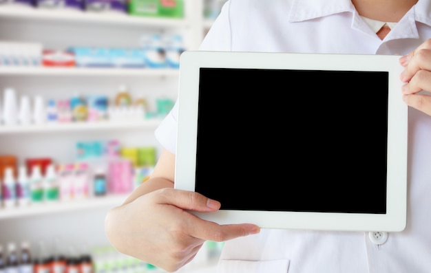 Farmacéutico en bata blanca mostrando tableta digital en blanco con estantes de medicamentos en la tienda de farmacia