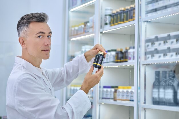 Farmacêutico de meia-idade calmo com um frasco de remédio de plástico lacrado em frente à vitrine da farmácia