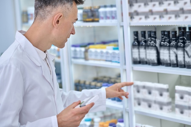 Farmacéutico experimentado con una túnica blanca centrado en buscar el medicamento adecuado en la vitrina de la farmacia.