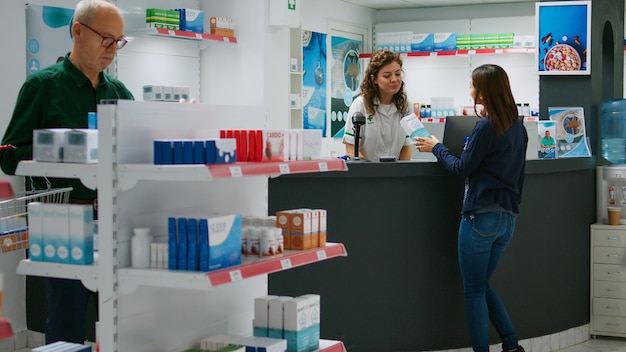 Farmacêutico feminino explicando produtos de saúde para cliente asiático no balcão da farmácia, falando sobre medicamentos prescritos e suplementos. Segurando o pacote de drogas na mesa da farmácia.