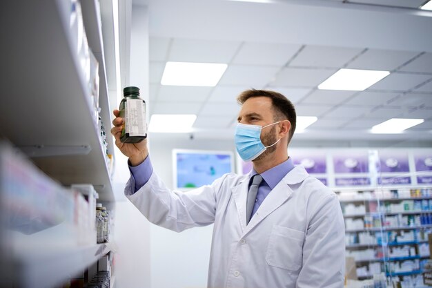 Farmacéutico con mascarilla y tomando vitaminas del estante en la farmacia durante la pandemia covid19