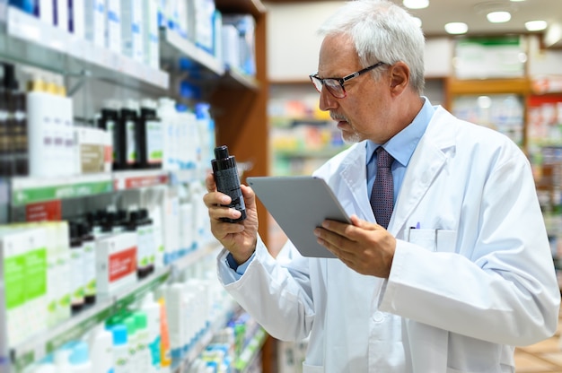 Farmacéutico senior buscando un producto en un estante y usando una tableta