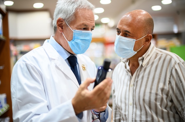 Foto farmacéutico senior que trata con un cliente, ambos con máscaras debido al coronavirus