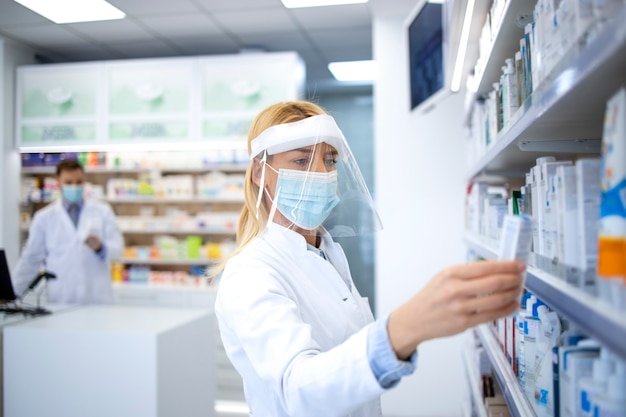 Farmacéutico de sexo femenino con protector facial y bata blanca con medicamento en la farmacia durante la pandemia del virus covid-19.