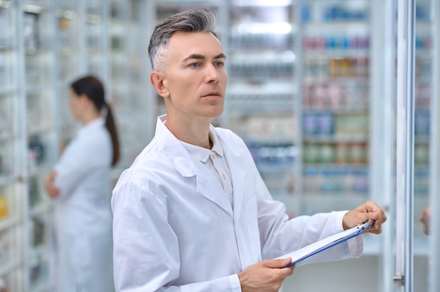 Farmacia, contabilidad. Hombre adulto en bata de laboratorio con carpeta mirando atentamente estantes con medicamentos y empleada en la parte trasera de la farmacia