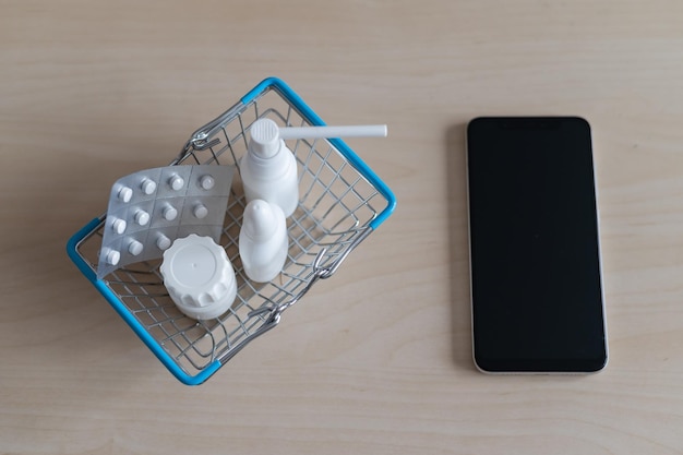 Farmacia en línea Blister con pastillas frasco de aerosol para la garganta con cápsulas en una mini cesta de la compra junto a un teléfono móvil con una aplicación de pantalla negra para comprar medicamentos