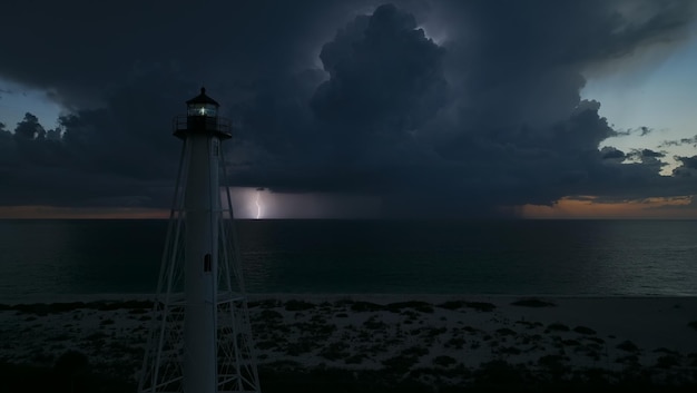 Faro alto blanco en la orilla del mar con luz parpadeante en la noche tormentosa para la navegación de embarcaciones comerciales Tormenta con relámpagos sobre el agua del océano que representa un peligro para los barcos