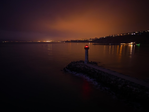 Farol à beira-mar com luz vermelha à noite contra o fundo das luzes dos navios