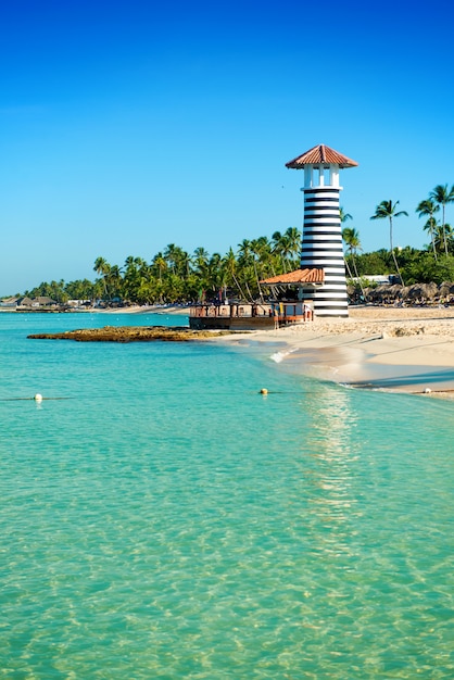 Farol listrado na costa arenosa com palmeiras. Águas claras do mar do Caribe.