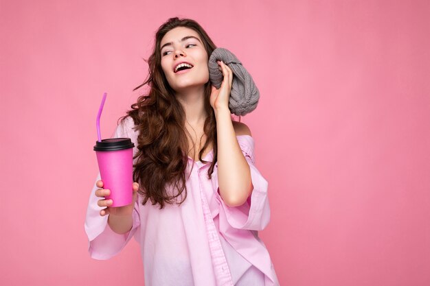 Fascinante jovem positiva morena encaracolada mulher vestindo camisa rosa e chapéu cinza isolado sobre o fundo rosa, segurando a xícara de café de papel para maquete de beber e se divertir olhando para o lado.