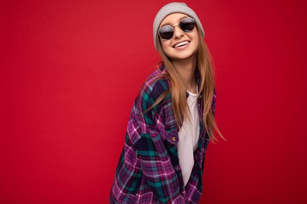 Fascinante mulher loira morena jovem e feliz isolada sobre um fundo vermelho, vestindo camisa casual roxa, chapéu cinza e óculos de sol elegantes, olhando para a câmera e sorrindo