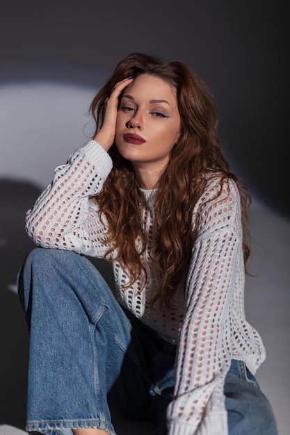 Fashion frisches schönes Model Mädchen in einem modischen weißen Pullover mit Jeans sitzt auf dem Boden im Studio auf einem dunklen Hintergrund Chic Lady