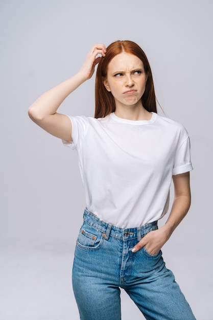 Fassungslos verwirrte junge Frau mit T-Shirt und Jeanshosen, die sich mit der Hand am Kopf kratzen