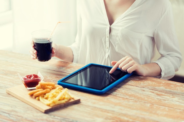 fast food, pessoas, tecnologia e conceito de dieta - close-up da mulher com computador tablet pc comendo batatas fritas com ketchup, anéis de lula fritos e cola na mesa de madeira