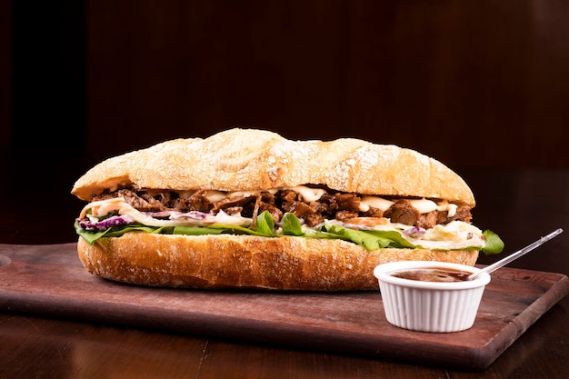 Fast-Food-Rinderbrustsandwich mit Rucola und Krautsalat auf Baguettebrot auf Holzbrett mit Barbecue-Sauce dunklem Hintergrund von vorne