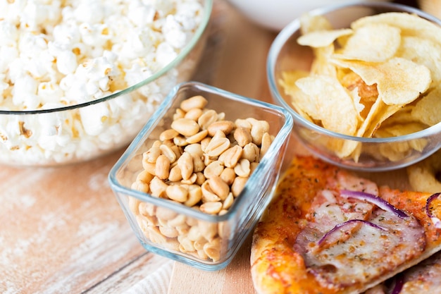 fast food und ungesundes essen konzept - nahaufnahme von erdnüssen in glasschale, kartoffelchips, pizza und popcorn auf holztisch